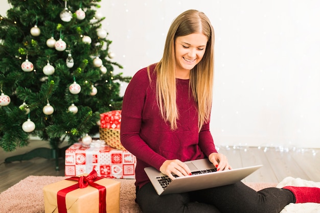 Señora sonriente con el ordenador portátil cerca de cajas de regalo y árbol de Navidad
