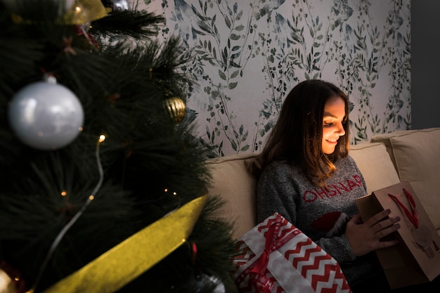 Señora sonriente con el paquete del regalo en el settee cerca del árbol de navidad