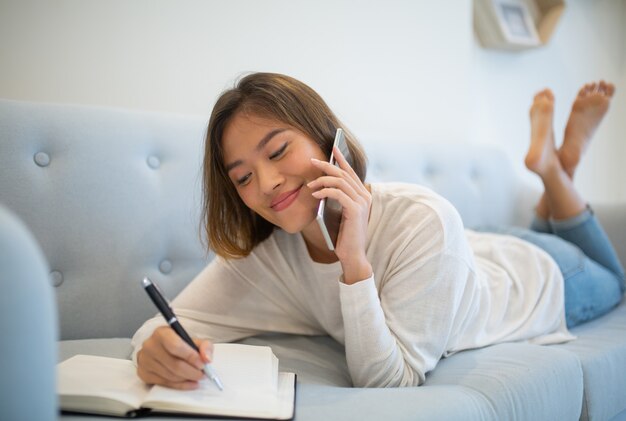 Señora sonriente que hace notas y que habla en el teléfono en casa