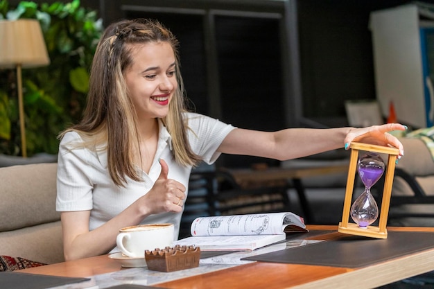 Señora sonriente que sostiene el reloj de arena y el pulgar del gesto para arriba