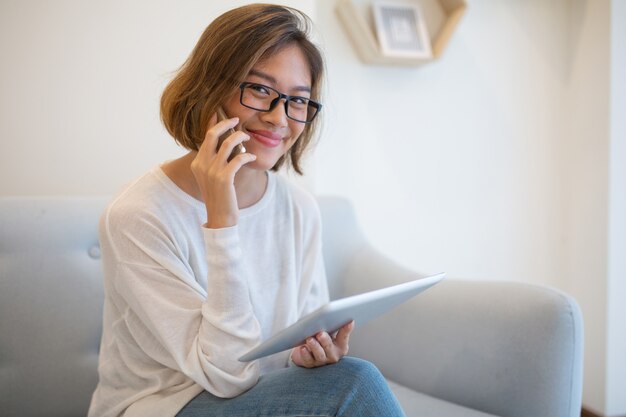 Señora sonriente que sostiene la tableta y que habla en el teléfono en el sofá