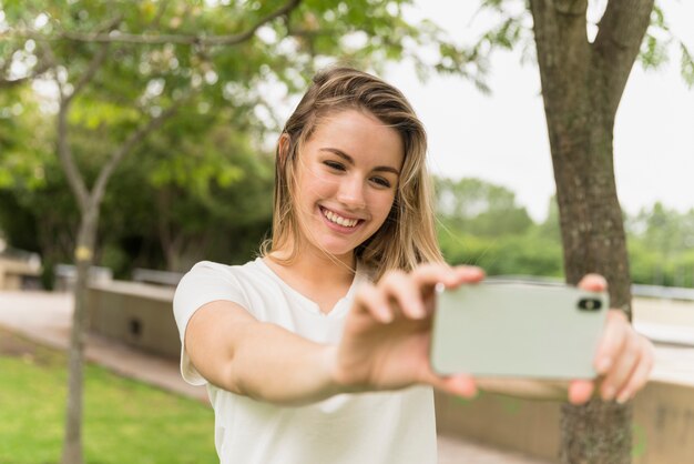 Señora sonriente que toma el selfie en el teléfono móvil en parque
