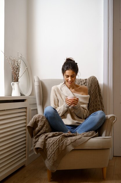 Señora sonriente en ropa de moda elegante está sentada en un sillón con una taza de té.