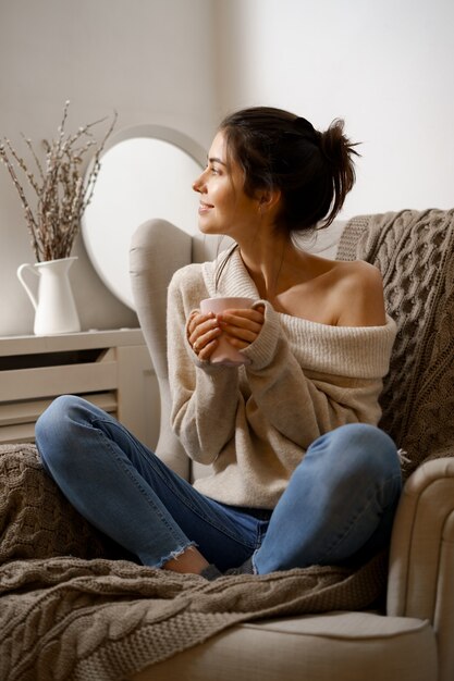 Señora sonriente en ropa de moda elegante está sentada en un sillón con una taza de té.