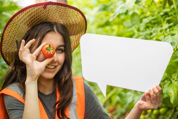 Señora sonriente sosteniendo tablero de ideas y tomate en el invernadero