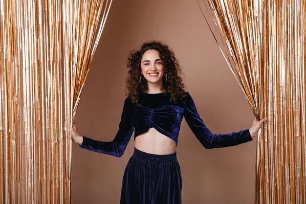 Señora sonriente en traje azul oscuro posando sobre fondo de cortinas doradas