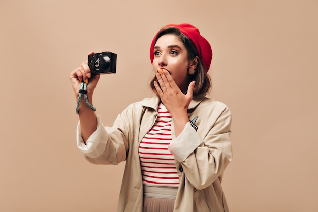 Señora sorprendida en boina roja y abrigo de moda sostiene la cámara. Mujer linda en suéter de rayas y abrigo ligero posando sobre fondo aislado.