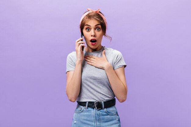 Señora sorprendida en traje gris charlando por teléfono. Moda mujer fresca en bandana rosa y falda de mezclilla con cinturón negro mirando a la cámara.