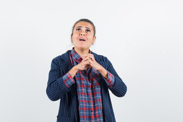 Señora sosteniendo los dedos entrelazados en camisa, chaqueta y mirando soñadora, vista frontal.