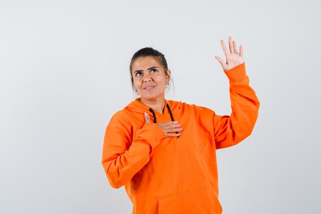 Señora en sudadera con capucha naranja agitando la mano para saludar y mirar alegre