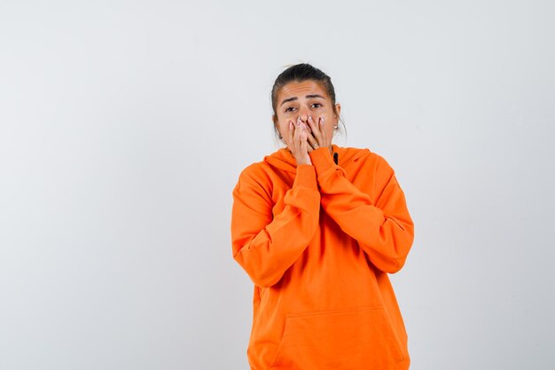 Señora en sudadera con capucha naranja manteniendo las manos en la boca y mirando emocionada