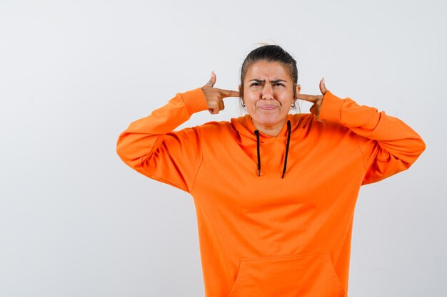 Señora en sudadera con capucha naranja tapando las orejas con los dedos y mirando irritada