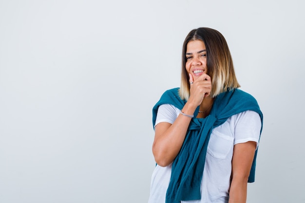 Señora con suéter atado en camiseta blanca con la mano en la barbilla y mirando alegre, vista frontal.