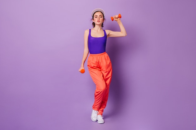 Señora en top deportivo morado y pantalón naranja haciendo ejercicios para manos con mancuernas rojas