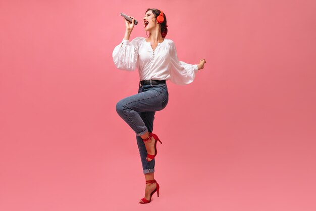 Señora en traje elegante canta emocionalmente en micrófono sobre fondo rosa. Hermosa mujer joven en auriculares rojos posando.