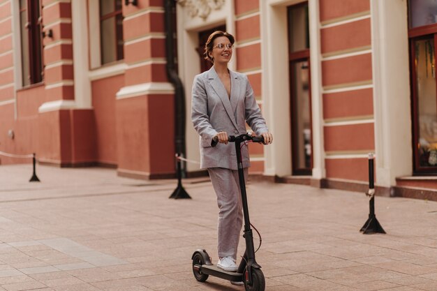 Señora en traje elegante divirtiéndose y montando scooter. Mujer muy feliz en chaqueta gris de gran tamaño y pantalones sonriendo y disfrutando de la vista de la ciudad