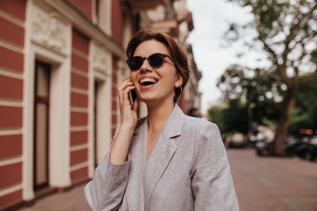 Señora en traje gris sonriendo y hablando por teléfono afuera. Feliz emocionada mujer de pelo corto en chaqueta de gran tamaño riendo y caminando por la ciudad