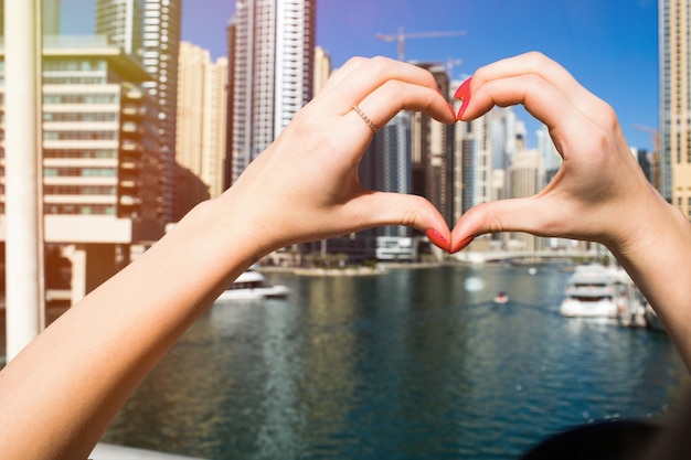 Señora con las uñas rojas demuestra el corazón con sus dedos antes de rascacielos de Dubai