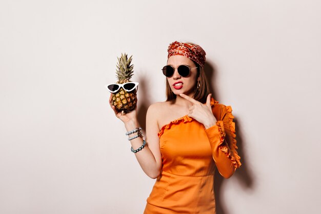 Señora con vestido naranja y gafas de sol está posando pensativamente y sosteniendo piña en un espacio aislado.