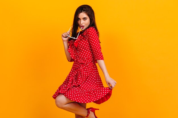 Señora en vestido rojo de lunares posando coquetamente en la pared amarilla, quitándose las gafas de sol.