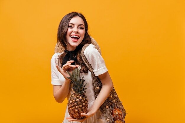 Señora en vestido de verano de algodón se ríe y posa con piña y bolsa ecológica sobre fondo naranja.