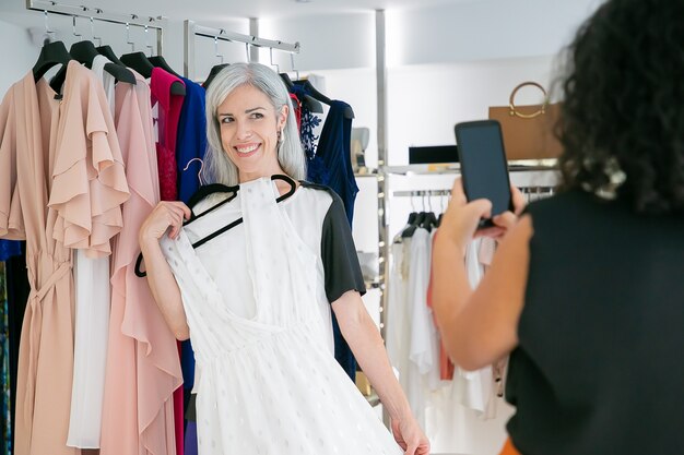 Señoras alegres disfrutando juntos de compras en la tienda de moda, sosteniendo el vestido y tomando fotografías en el teléfono celular. Consumismo o concepto de compras