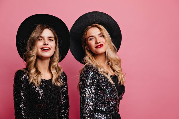 Señoras blancas alegre con sombreros posando en la pared rosada. Retrato de interior de chicas caucásicas seguras con accesorios glamorosos.