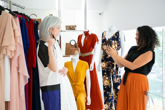 Señoras felices disfrutando juntos de compras en la tienda de moda, eligiendo el vestido y tomando fotografías en el teléfono inteligente. Vista lateral. Consumismo o concepto de compras