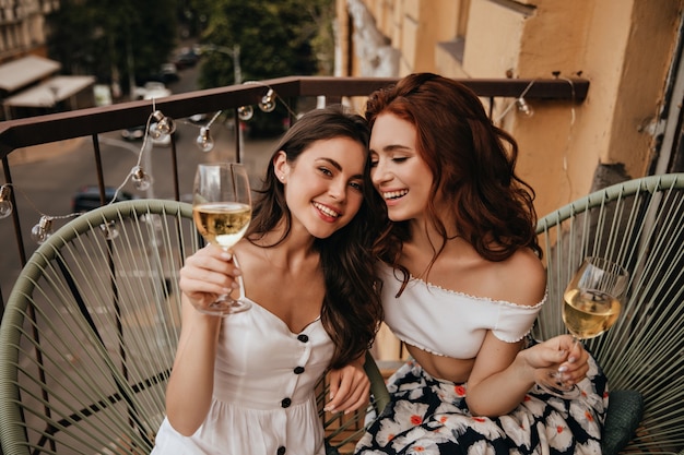 Señoras felices con trajes elegantes disfrutan de vino blanco