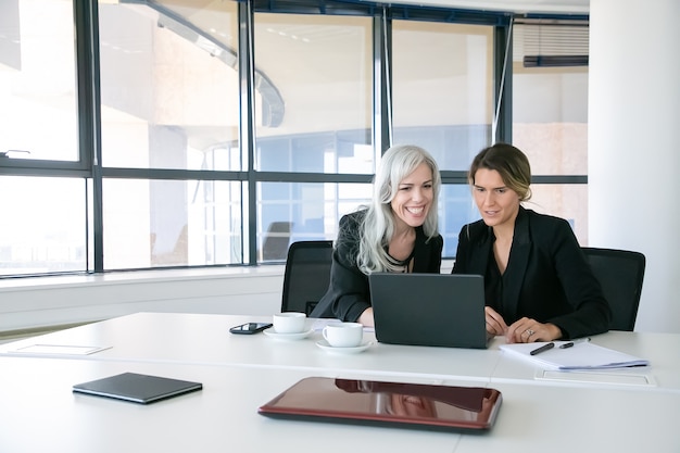 Señoras de negocios alegres mirando la pantalla del portátil, hablando y sonriendo mientras está sentado a la mesa con tazas de café en la oficina. Concepto de comunicación y trabajo en equipo