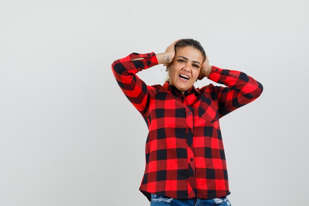 Señorita agarrando la cabeza con las manos en camisa a cuadros, pantalones cortos y mirando alegre.