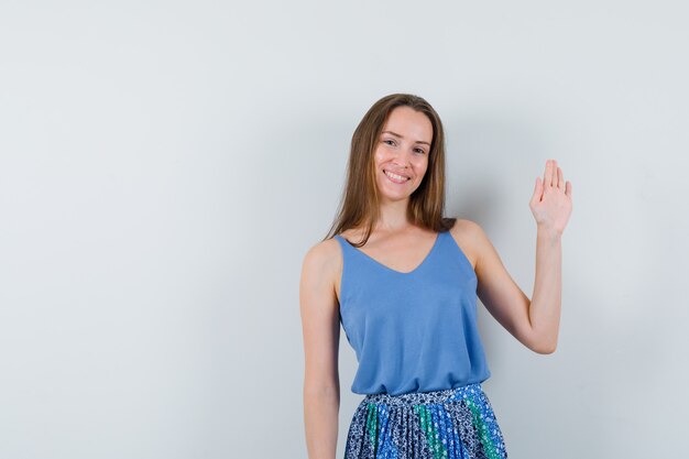 Señorita agitando la mano para decir adiós en camiseta, falda y mirando alegre, vista frontal.