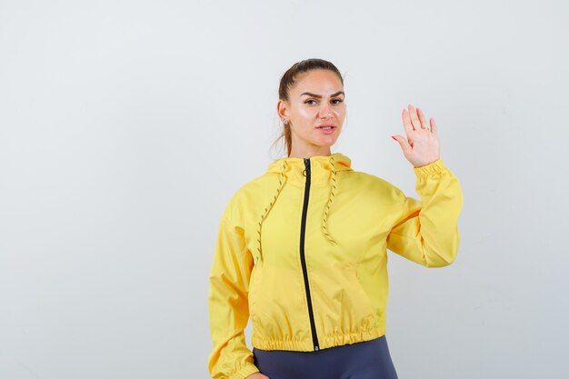 Señorita agitando la mano para decir adiós en chaqueta amarilla y mirando confiado. vista frontal.