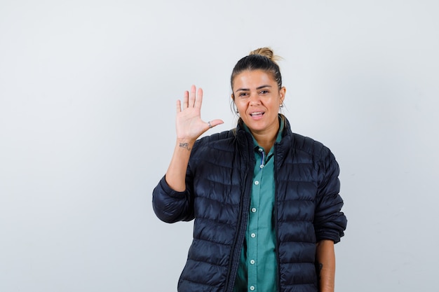 Señorita agitando la mano para saludar en camisa, chaqueta acolchada y mirando alegre, vista frontal.