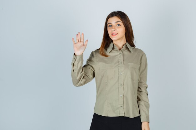 Señorita agitando la mano para saludar en camisa, falda y mirando alegre, vista frontal.