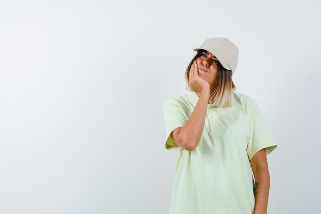 Señorita apoyando la barbilla en la mano en camiseta, gorra y mirando pensativo, vista frontal.