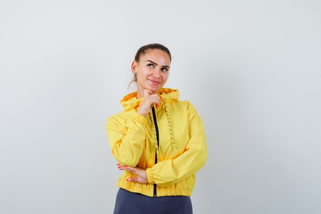 Señorita apoyando la barbilla en la mano con chaqueta amarilla y buscando pacífica. vista frontal.