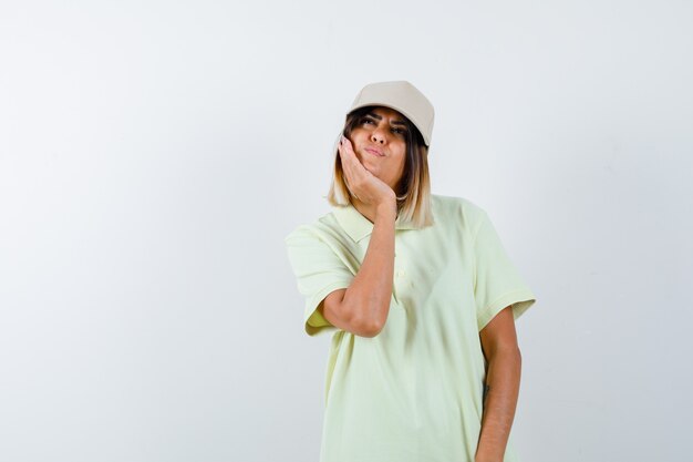 Señorita apoyando la barbilla en su mano en camiseta, gorra y mirando pensativo, vista frontal.