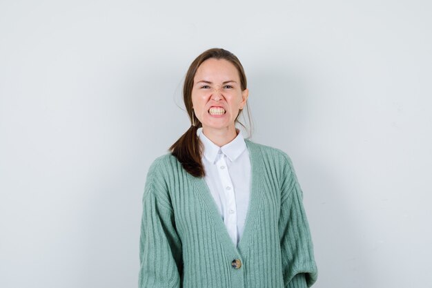 Señorita apretando los dientes en blusa, chaqueta de punto y mirando furiosa, vista frontal.