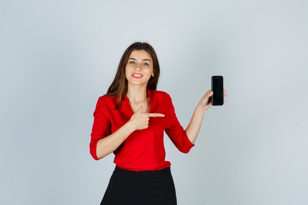 Señorita apuntando al teléfono móvil en blusa roja, falda y mirando alegre