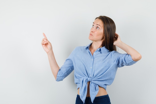 Señorita apuntando hacia arriba con camisa azul, pantalones y mirando vacilante.