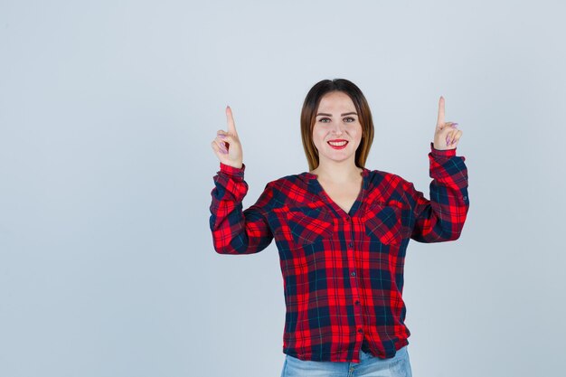 Señorita apuntando hacia arriba en camisa a cuadros, jeans y mirando complacido. vista frontal.