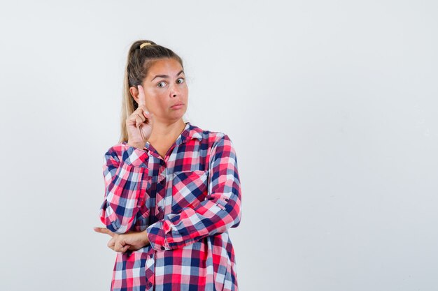 Señorita apuntando hacia arriba con camisa a cuadros y mirando inteligente, vista frontal.