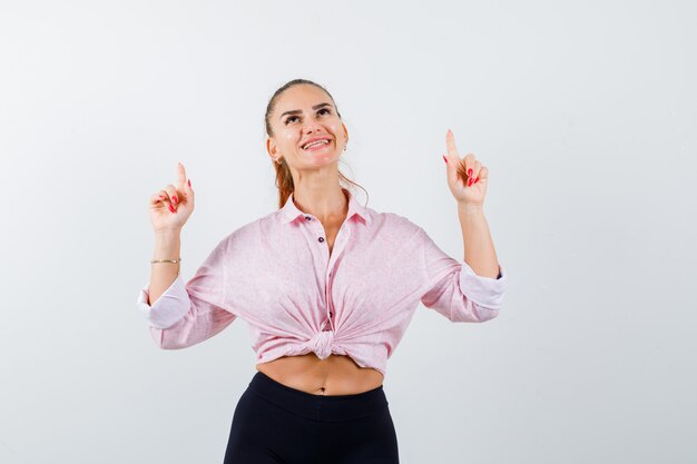 Señorita apuntando hacia arriba en camisa, pantalones y mirando alegre. vista frontal.