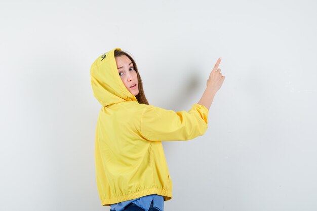 Señorita apuntando hacia arriba en camiseta, chaqueta y mirando vacilante.