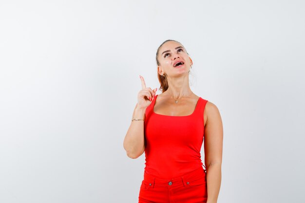 Señorita apuntando hacia arriba en camiseta roja, pantalón rojo y mirando alegre, vista frontal.