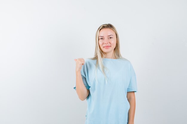 Señorita apuntando hacia atrás con el pulgar en camiseta y mirando confiado, vista frontal.