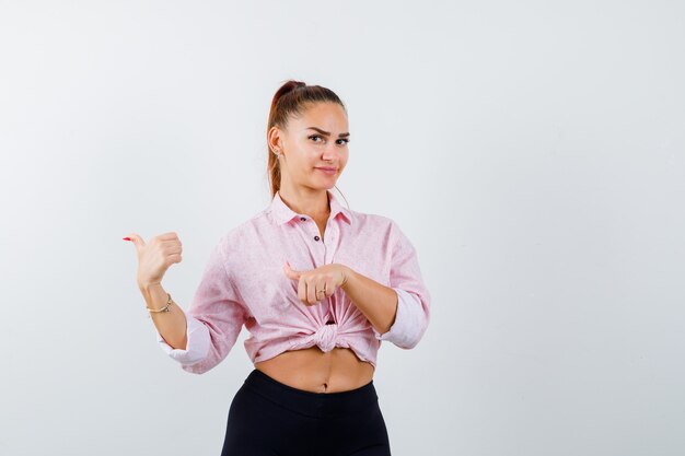 Señorita apuntando hacia atrás con los pulgares en camisa, pantalones y mirando confiado, vista frontal.