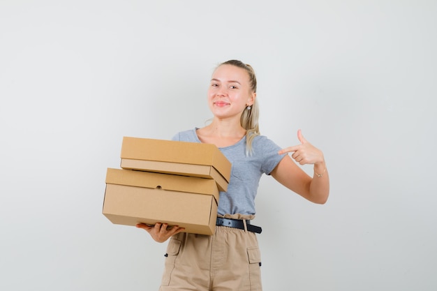 Señorita apuntando a cajas de cartón en camiseta y pantalón y mirando alegre