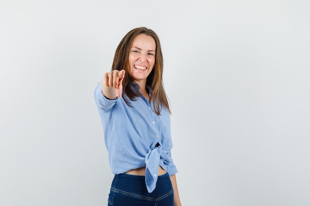 Señorita apuntando a la cámara en camisa azul, pantalones y mirando feliz